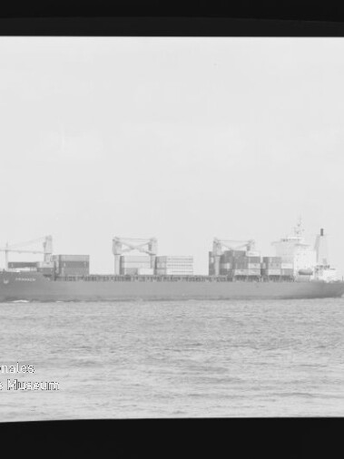 Seitenansicht eines Containerschiffs mit aufgestapelten Ladecontainern und mehreren Bordkränen auf ruhiger See vor hellem Himmel, links unten sichtbar das Wasserzeichen des Internationalen Maritimen Museums Hamburg.
