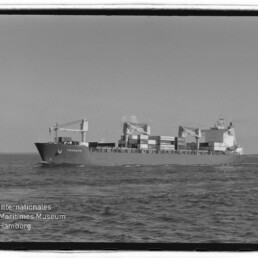 Schwarzweiß-Aufnahme eines Containerschiffs auf ruhiger See, Seitenansicht mit drei Ladekränen und gestapelten Containern; unten links das Logo des Internationalen Maritimen Museums Hamburg.