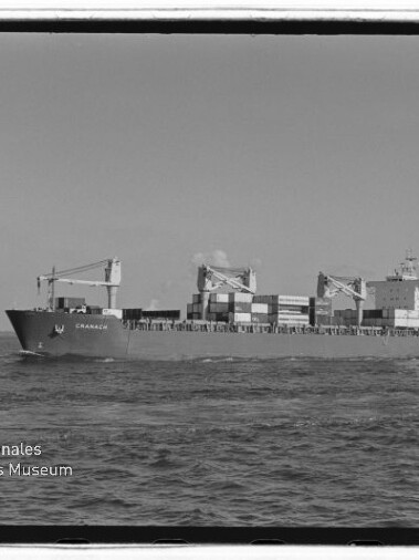 Schwarzweiß-Aufnahme eines Containerschiffs auf ruhiger See, Seitenansicht mit drei Ladekränen und gestapelten Containern; unten links das Logo des Internationalen Maritimen Museums Hamburg.