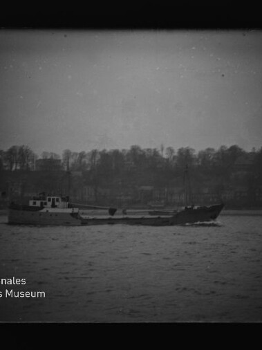 Schwarzweiß-Foto eines niedrigen Frachtschiffs, das auf ruhigem Wasser vor einer baumbestandenen Uferlinie mit Gebäudesilhouetten fährt; leicht körnig, mit Museumslogo unten links.