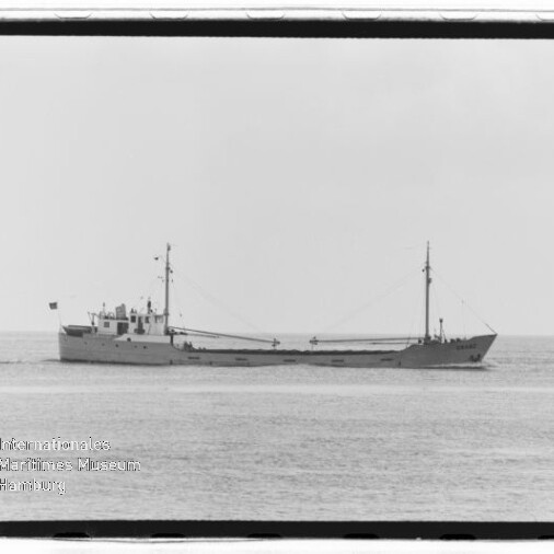 Schwarzweiß-Seitenaufnahme eines langen Frachtschiffs auf ruhiger See mit Brückenaufbau mittschiffs, zwei Masten und kleiner Flagge, weiter flacher Horizont im Hintergrund.