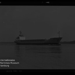 Schwarzweißaufnahme eines Frachtschiffs in Seitenansicht, das ruhig über ruhiges Wasser vor einem flachen Horizont fährt; links unten sichtbar Logo und Schriftzug des Internationalen Maritimen Museums Hamburg.