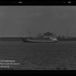 Schwarzweißaufnahme eines Frachtschiffs, das auf ruhiger See von links nach rechts fährt, im Hintergrund eine Industriehafen-Kulisse mit Fabrikgebäuden, Schornsteinen und Strommasten, links unten das Logo des Internationalen Maritimen Museums Hamburg.