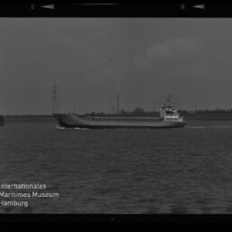 Schwarzweiß-Foto eines Frachtschiffs, das auf ruhigem Wasser an einer flachen Küste mit Hochspannungsleitungen vorbeifährt; im Bildrand ist ein Filmrahmen sichtbar und unten links das Logo sowie der Schriftzug des Internationalen Maritimen Museums Hamburg.