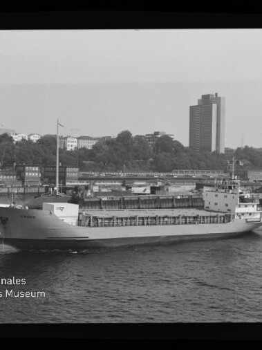 Schwarz-weiß-Foto eines Frachtschiffs, das am Kai eines Hafens vorbeifährt; im Hintergrund Uferbebauung mit Bäumen und einem hohen Büroturm, unten links das Wasserzeichen des Internationalen Maritimen Museums Hamburg.