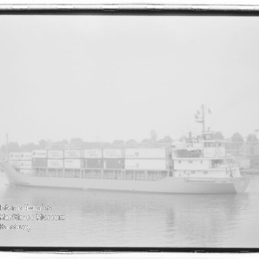 Frachtschiff mit mehreren übereinander gestapelten Containern fährt auf ruhigem Wasser vor einer nebligen Uferlinie mit Gebäuden und Bäumen.