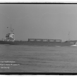 Schwarzweißaufnahme eines Frachtschiffs auf ruhiger See mit mehreren auf dem Deck gestapelten Containern, auf denen mehrfach „CAST“ steht, unten links Logo und Schriftzug des Internationalen Maritimen Museums Hamburg