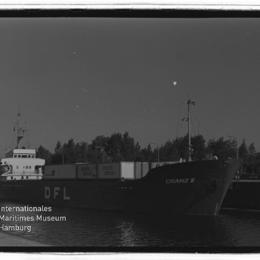 Schwarzweißfoto eines Frachtschiffs mit der Beschriftung DFL und dem Namen CRANZ II, beladen mit Containern am Kai vor einer Baumreihe; links unten das Logo und Wasserzeichen des Internationalen Maritimen Museums Hamburg.