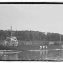 Schwarzweißaufnahme eines Frachtschiffs mit großen Buchstaben DFL am Rumpf, beladen mit Containern und fahrend auf einem ruhigen Kanal vor einem bewaldeten Ufer, mit Bug- und Heckwellen.