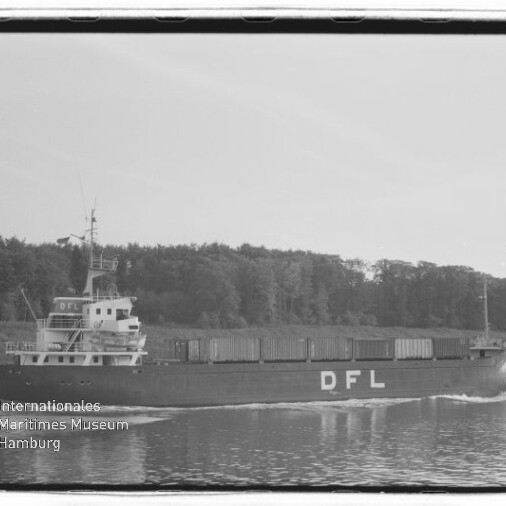 Schwarzweißaufnahme eines Frachtschiffs mit großen Buchstaben DFL am Rumpf, beladen mit Containern und fahrend auf einem ruhigen Kanal vor einem bewaldeten Ufer, mit Bug- und Heckwellen.