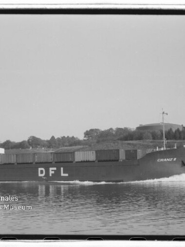 Schwarzweißfoto eines Frachtschiffs mit der Bordaufschrift DFL und dem Namen CRANZ II, beladen mit Containern und fahrend auf einem Kanal, im Hintergrund Ufer mit Bäumen und Tankanlagen.
