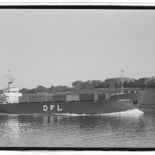 Schwarzweißfoto eines Frachtschiffs mit der Bordaufschrift DFL und dem Namen CRANZ II, beladen mit Containern und fahrend auf einem Kanal, im Hintergrund Ufer mit Bäumen und Tankanlagen.
