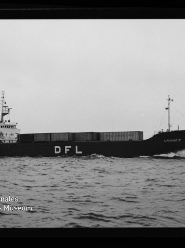 Schwarzweiß-Seitenaufnahme eines Frachtschiffs mit aufgestapelten Containern und großer weißer Aufschrift „DFL“ am Rumpf, das bei leicht bewegter See unter grauem Himmel fährt; Museumslogo unten links sichtbar.