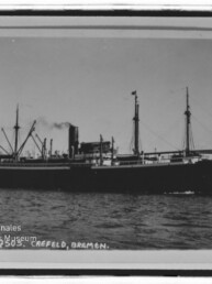 Schwarzweiß-Foto eines großen Frachtdampfers (S.S. CREFELD) mit rauchendem Schornstein, mehreren Masten und Ladebäumen, in Fahrt auf ruhiger See; links unten Stempel des Internationalen Maritimen Museums Hamburg.