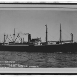 Schwarzweiß-Foto eines großen Frachtdampfers (S.S. CREFELD) mit rauchendem Schornstein, mehreren Masten und Ladebäumen, in Fahrt auf ruhiger See; links unten Stempel des Internationalen Maritimen Museums Hamburg.