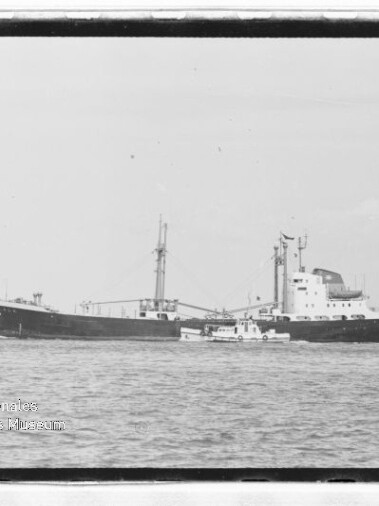 Schwarzweißaufnahme eines großen Frachtschiffs mit Mast und achterlichem Aufbau, neben dem ein kleines Begleitboot auf ruhiger See vor klarem Horizont fährt.