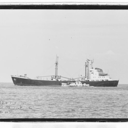 Schwarzweißaufnahme eines großen Frachtschiffs mit Mast und achterlichem Aufbau, neben dem ein kleines Begleitboot auf ruhiger See vor klarem Horizont fährt.