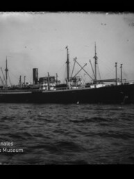Schwarz-weiß-Fotografie eines großen Dampffrachters mit hohem Schornstein, mehreren Masten und Ladebäumen auf leicht bewegter See vor einem Hafen, mit Wasserzeichen des Internationalen Maritimen Museums Hamburg in der linken unteren Ecke.