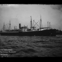 Schwarz-weiß-Fotografie eines großen Dampffrachters mit hohem Schornstein, mehreren Masten und Ladebäumen auf leicht bewegter See vor einem Hafen, mit Wasserzeichen des Internationalen Maritimen Museums Hamburg in der linken unteren Ecke.