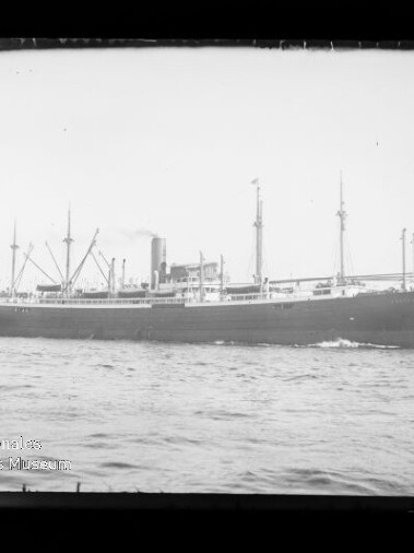 Seitliche Schwarzweißaufnahme eines großen Frachtdampfers mit mehreren Ladebäumen und einem zentralen Schornstein, der auf ruhiger See entlangfährt; unten links sichtbar das Wasserzeichen des Internationalen Maritimen Museums Hamburg.