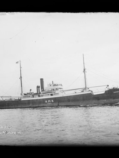 Schwarzweißaufnahme eines Frachtdampfers mit zwei Masten und einem Schornstein, die Aufschrift H.M.G. am Rumpf sichtbar, fahrend auf ruhigem Wasser vor flachem Horizont; Museumslogo unten links.