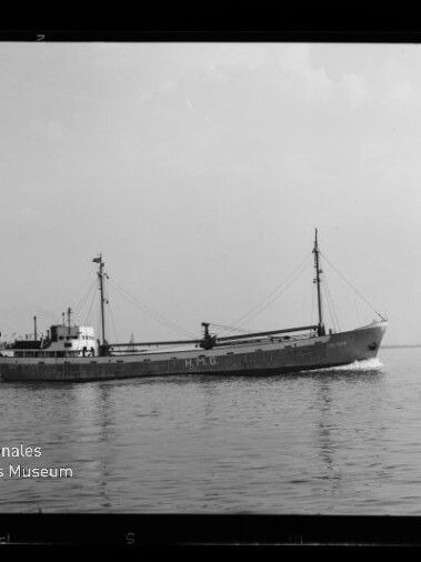 Schwarzweiß-Foto eines Frachtschiffs mit zwei Masten und der Aufschrift HMG auf dem Rumpf, das ruhig über das Meer fährt; links im Hintergrund ein kleines Segelboot und unten links das Logo des Internationalen Maritimen Museums Hamburg.