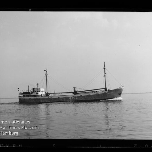 Schwarzweiß-Foto eines Frachtschiffs mit zwei Masten und der Aufschrift HMG auf dem Rumpf, das ruhig über das Meer fährt; links im Hintergrund ein kleines Segelboot und unten links das Logo des Internationalen Maritimen Museums Hamburg.