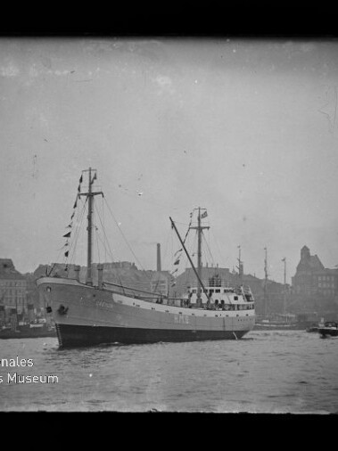 Schwarz-weiß-Foto eines Frachtschiffs mit zwei Masten und Signalflaggen im Hafen vor einer städtischen Uferkulisse, ein kleines Begleitboot neben dem Schiff und Wasserzeichen des Internationalen Maritimen Museums Hamburg.
