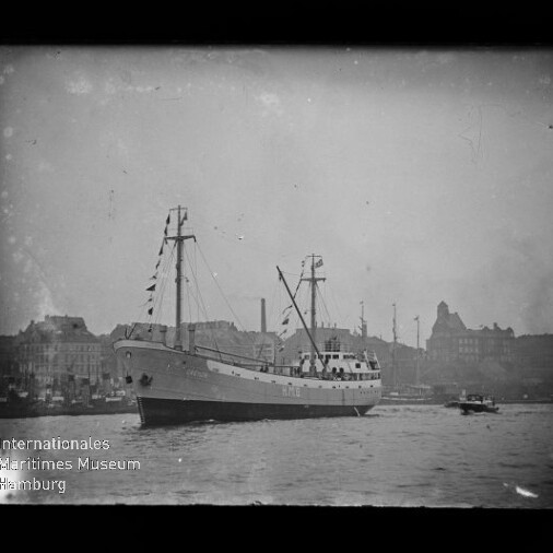 Schwarz-weiß-Foto eines Frachtschiffs mit zwei Masten und Signalflaggen im Hafen vor einer städtischen Uferkulisse, ein kleines Begleitboot neben dem Schiff und Wasserzeichen des Internationalen Maritimen Museums Hamburg.