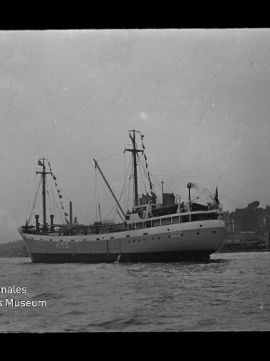 Schwarz-Weiß-Foto eines mittelgroßen Schiffes mit hellem Rumpf, zwei Masten und sichtbarer Takelage, vor einer Hafenpromenade mit Gebäuden im Hintergrund und Museumslogo unten links.
