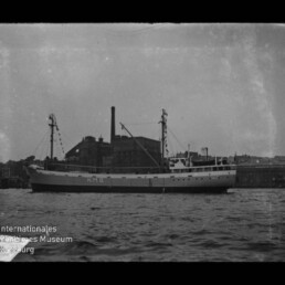 Schwarzweiß-Foto eines mittelgroßen Frachtschiffs mit der Aufschrift „M.F.G.“ am Rumpf, das im Hafen vor einer Industriekaianlage mit hohem Schornstein liegt; im Vordergrund ruhige See und unten links ein Museumswasserzeichen.