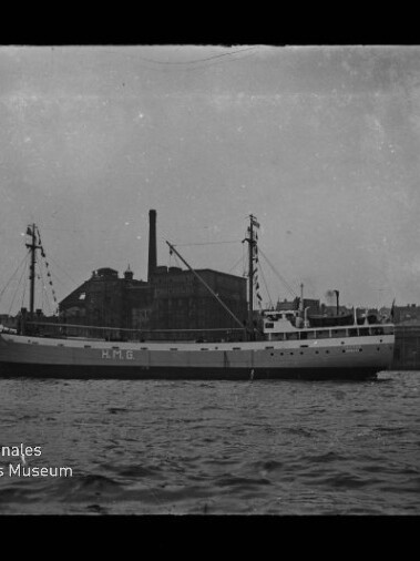 Schwarzweiß-Foto eines mittelgroßen Frachtschiffs mit der Aufschrift „M.F.G.“ am Rumpf, das im Hafen vor einer Industriekaianlage mit hohem Schornstein liegt; im Vordergrund ruhige See und unten links ein Museumswasserzeichen.