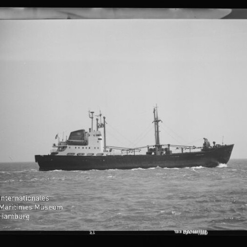 Schwarzweiß-Foto eines Frachtschiffs in Profil auf ruhiger See mit mittigem Aufbauten, zwei Masten und Ladegeschirr; unten links das Wasserzeichen Internationales Maritimes Museum Hamburg.