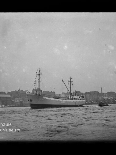 Schwarzweißaufnahme eines geschmückten Frachtschiffs mit zwei Masten, das im Hafen vor einer Stadtkulisse mit Kirchtürmen fährt; im Vordergrund unruhiges Wasser und kleine Begleitboote, links unten das Logo des Internationalen Maritimen Museums Hamburg.