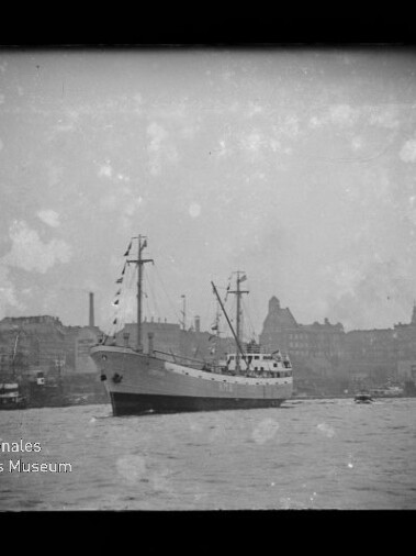 Schwarz-weiß-Foto eines mittelgroßen Frachtschiffs mit zwei Masten und Flaggen vor einer städtischen Hafen-Silhouette, Wasser im Vordergrund und sichtbare Alterungsflecken; Logo des Internationalen Maritimen Museums Hamburg unten links.