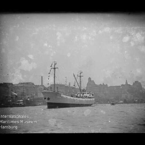 Schwarz-weiß-Foto eines mittelgroßen Frachtschiffs mit zwei Masten und Flaggen vor einer städtischen Hafen-Silhouette, Wasser im Vordergrund und sichtbare Alterungsflecken; Logo des Internationalen Maritimen Museums Hamburg unten links.
