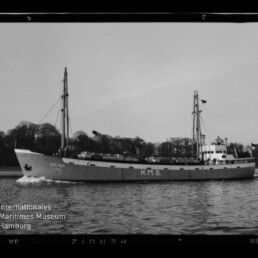 Schwarzweiß-Foto eines zweimastigen Frachtschiffs (Name 'CREMON') mit großer Aufschrift 'H.M.G.' am Rumpf, das mit Bugwelle auf ruhiger See vor einer baumbestandenen Küstenlinie fährt; links unten das Logo des Internationalen Maritimen Museums Hamburg.