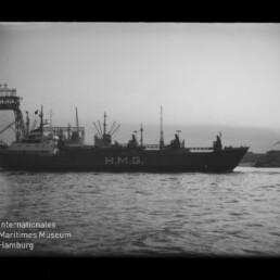Schwarz-weiß-Foto eines Frachtschiffs mit großer Aufschrift H.M.G. an der Bordwand, seitlich vor einem Industriehafen mit Kränen und Lagerbauten auf ruhigem Wasser, unten links Museum-Logo sichtbar