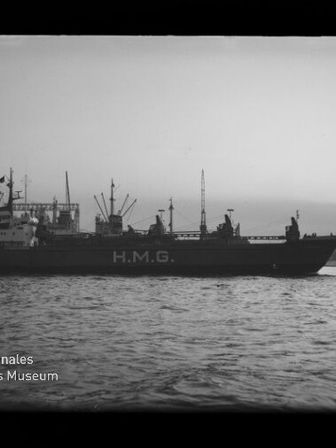 Schwarz-weiß-Foto eines Frachtschiffs mit großer Aufschrift H.M.G. an der Bordwand, seitlich vor einem Industriehafen mit Kränen und Lagerbauten auf ruhigem Wasser, unten links Museum-Logo sichtbar