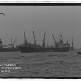 Schwarzweiß-Foto eines großen Frachtschiffs mit der Aufschrift HMG im Hafen, im Hintergrund mehrere Hafenkräne, rechts ein kleines Lotsenboot, unruhiges Wasser und Möwen am bewölkten Himmel; Museumslogo unten links.
