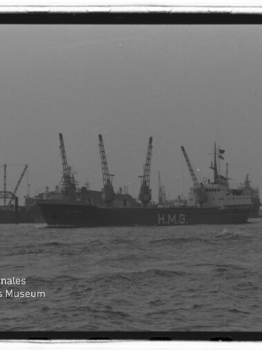Schwarzweiß-Foto eines großen Frachtschiffs mit der Aufschrift HMG im Hafen, im Hintergrund mehrere Hafenkräne, rechts ein kleines Lotsenboot, unruhiges Wasser und Möwen am bewölkten Himmel; Museumslogo unten links.