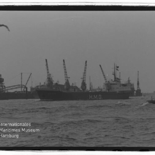 Schwarzweiß-Foto eines großen Frachtschiffs mit der Aufschrift HMG im Hafen, im Hintergrund mehrere Hafenkräne, rechts ein kleines Lotsenboot, unruhiges Wasser und Möwen am bewölkten Himmel; Museumslogo unten links.
