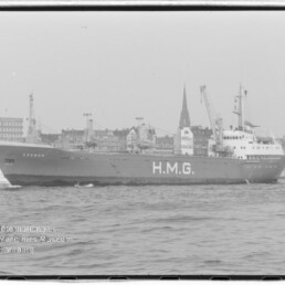 Schwarzweißaufnahme eines Frachtschiffs mit großem Schriftzug H.M.G. und dem Namen Cremon am Bug, fahrend auf der Elbe vor der Hamburger Uferfront mit Gebäuden und markanten Kirchtürmen im Hintergrund.