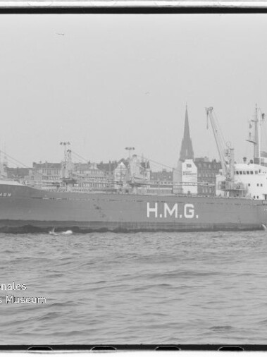 Schwarzweißaufnahme eines Frachtschiffs mit großem Schriftzug H.M.G. und dem Namen Cremon am Bug, fahrend auf der Elbe vor der Hamburger Uferfront mit Gebäuden und markanten Kirchtürmen im Hintergrund.