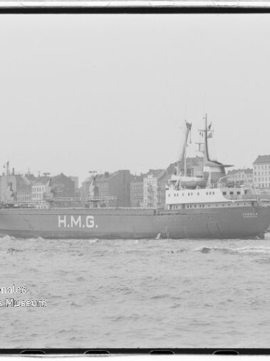 Schwarzweißaufnahme eines großen Schiffes mit der markanten Aufschrift H.M.G. auf dem Rumpf, das in Hafennähe vor einer städtischen Silhouette mit Kirchen- und Wohngebäuden vorbeifährt, rechts ein kleines Begleitboot, unten links Wasserzeichen des Internationalen Maritimen Museums Hamburg.