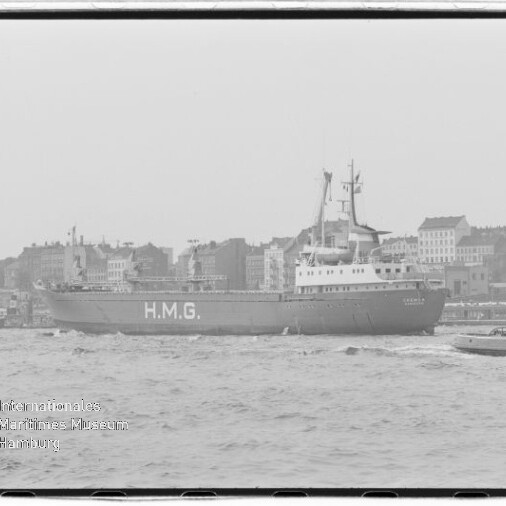 Schwarzweißaufnahme eines großen Schiffes mit der markanten Aufschrift H.M.G. auf dem Rumpf, das in Hafennähe vor einer städtischen Silhouette mit Kirchen- und Wohngebäuden vorbeifährt, rechts ein kleines Begleitboot, unten links Wasserzeichen des Internationalen Maritimen Museums Hamburg.