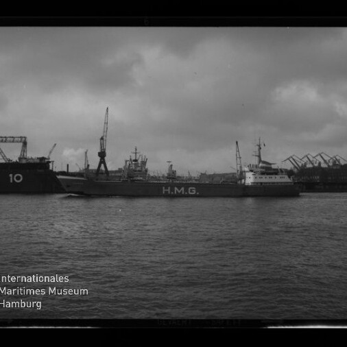 Schwarzweißaufnahme eines Frachtschiffs mit großer Aufschrift H.M.G., das auf dem Wasser vor einem Industriehafen mit mehreren Kränen und bewölktem Himmel fährt; unten links das Logo des Internationalen Maritimen Museums Hamburg.