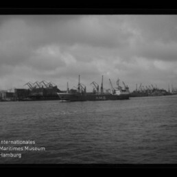 Schwarzweiß-Foto eines Frachtschiffs mit der Aufschrift H.M.G. auf dem Wasser vor einem Industriehafen mit zahlreichen Hafenkränen und einem bedeckten Himmel, unten links das Logo des Internationalen Maritimen Museums Hamburg