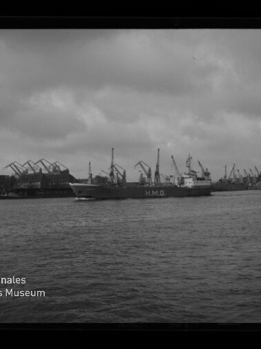 Schwarzweiß-Foto eines Frachtschiffs mit der Aufschrift H.M.G. auf dem Wasser vor einem Industriehafen mit zahlreichen Hafenkränen und einem bedeckten Himmel, unten links das Logo des Internationalen Maritimen Museums Hamburg
