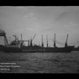 Schwarzweißaufnahme eines Frachtschiffs mit großer Aufschrift U.M.G. am Kai, im Vordergrund bewegte Wasseroberfläche und ein kleines Begleitboot, im Hintergrund mehrere hohe Hafenkräne; Museumslogo unten links.
